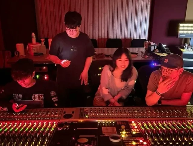 Musicians gathered around a mixing console in a red-lit studio.